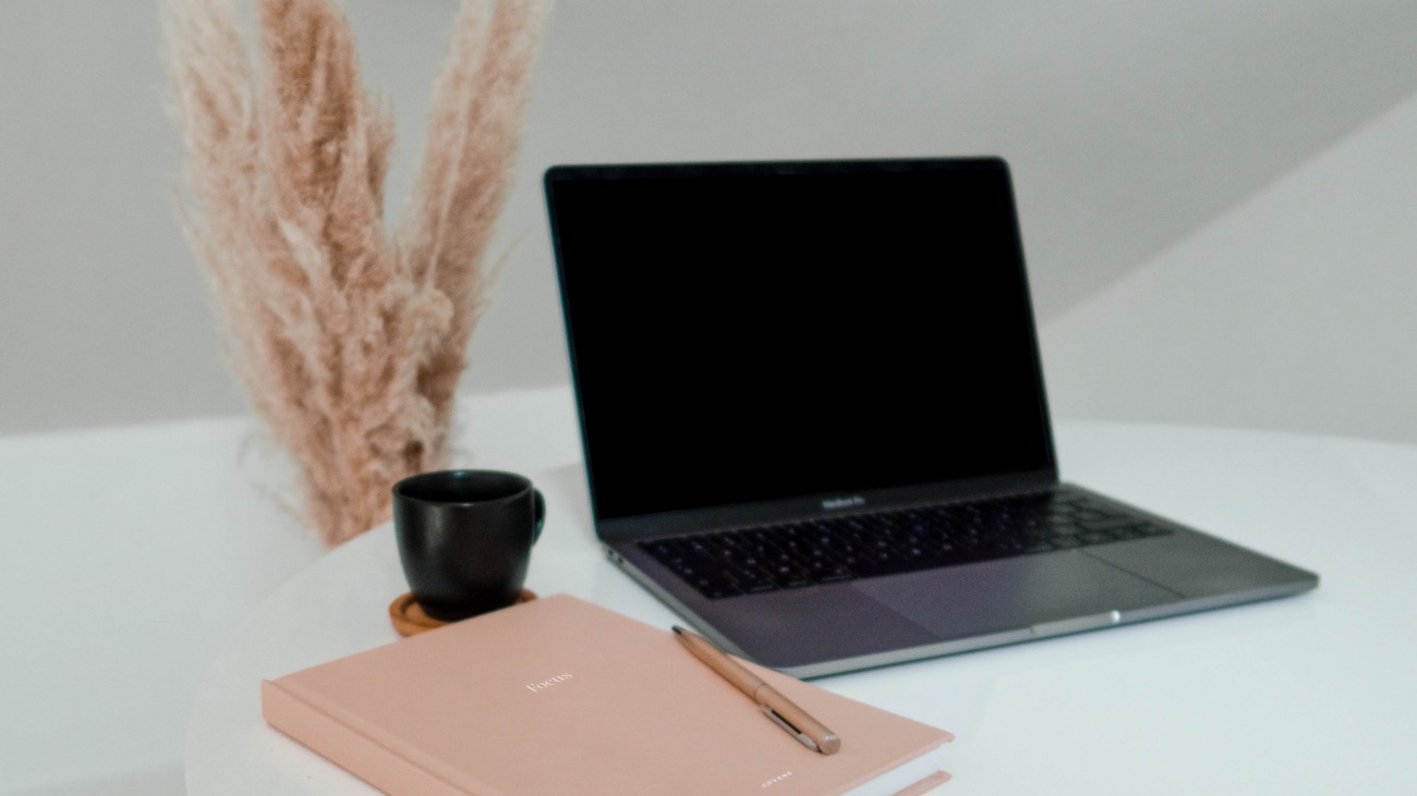 a laptop with a pink notebook and pampas grass in the background